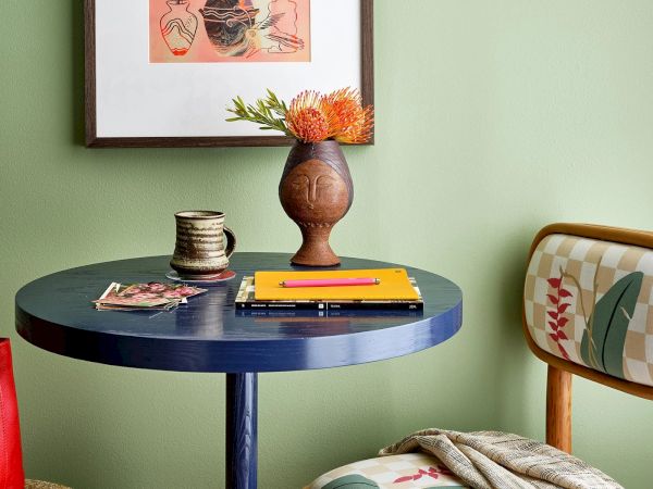 The image features a small round blue table with a vase of flowers, a mug, a notebook with a pen, a picture frame, and a chair with a patterned cushion.