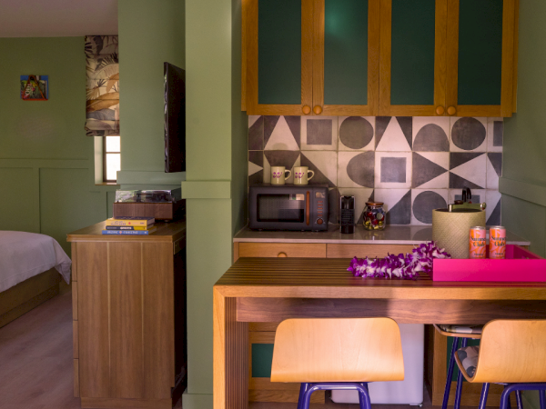 Una cocina compacta y una zona de escritorio en una habitaci&oacute;n pintada de verde, con una isla de madera, dos sillas claras, estanter&iacute;as abiertas, un microondas y una bandeja rosa; parte de una cama y una peque&ntilde;a ventana son visibles al fondo.