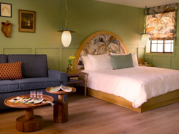 A cozy bedroom with a double bed, a blue couch, wooden tables, wall art, and green walls. The room is well-lit with a window and hanging lights.