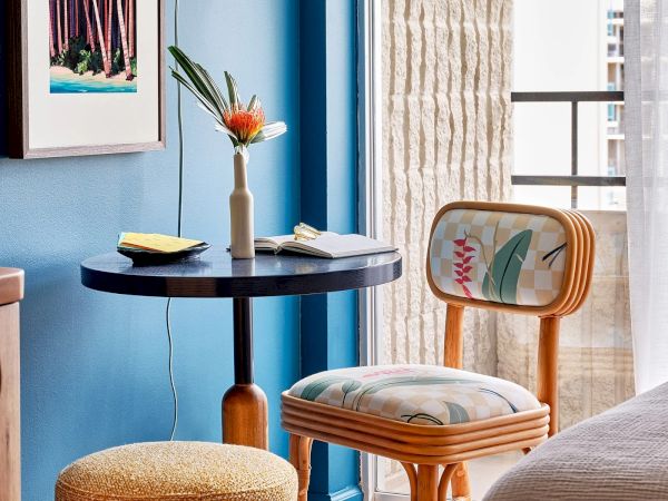 A cozy corner with a small round table, floral chair, ottoman, decorative vase with a flower, and an artwork on a blue wall.