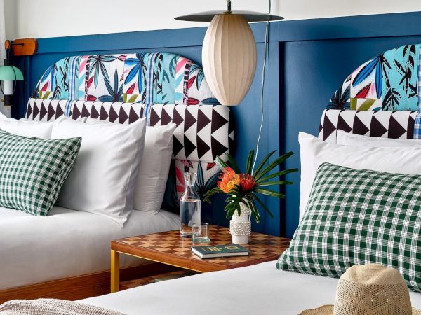 A hotel room features two double beds with colorful headboards, green checkered pillows, a side table with a lamp, flowers, and a hat.