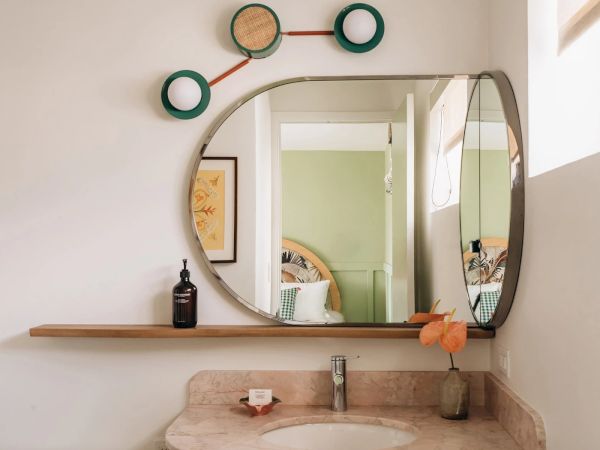 This image displays a modern bathroom with a circular and rectangular mirror, decorative wall art, a sink, and a shelf with a bottle and plant.