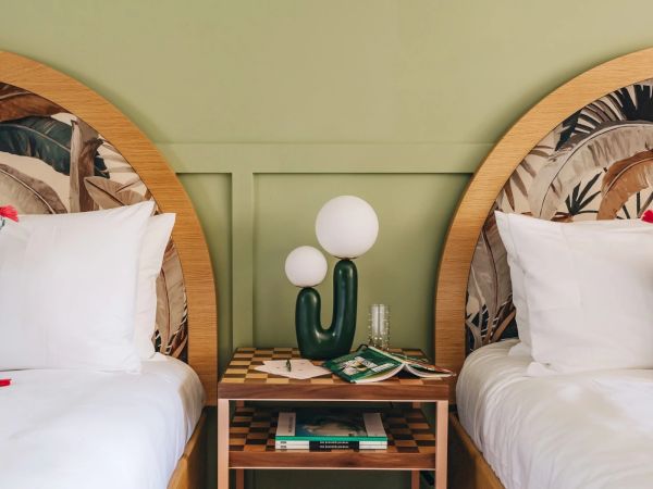 The image shows a cozy bedroom with two single beds, a green cactus-shaped lamp on a nightstand between them, and patterned headboards.