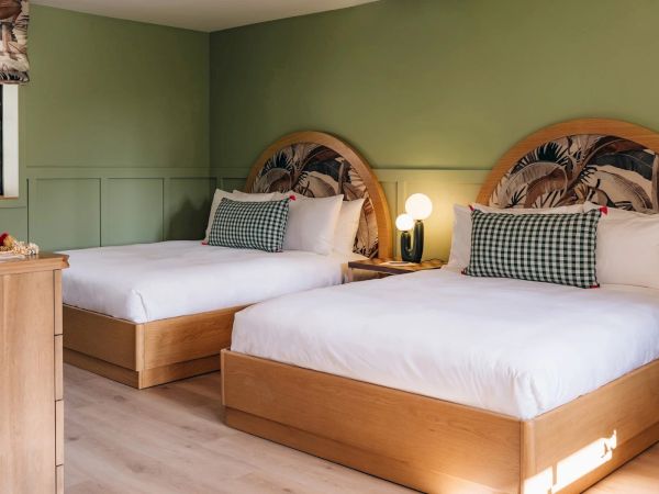 A cozy bedroom with two neatly made beds, wooden frames, checkered pillows, and a bedside table with a round lamp, all against green walls.