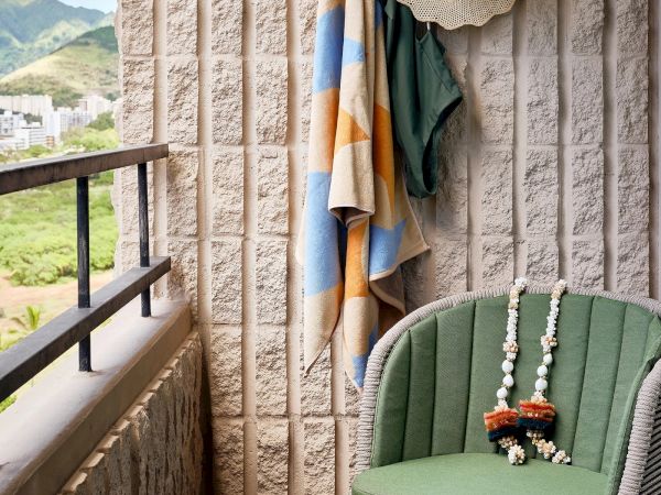 A small balcony with a green chair, bordered by a railing, has colorful towels, a hat hung on the wall, and a necklace draped over the chair.