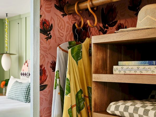 A cozy room with a bed, books on a shelf, a hat, and clothing hanging. The wallpaper features a floral pattern. The room has a warm and inviting vibe.