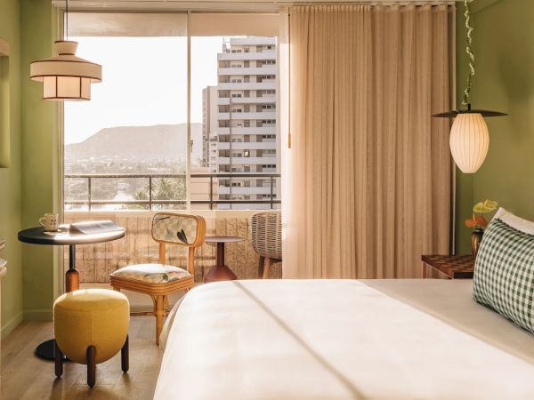 A cozy room with a bed, stylish lamps, a small table, chairs, and a balcony with a view of a distant mountain and nearby buildings, is seen.