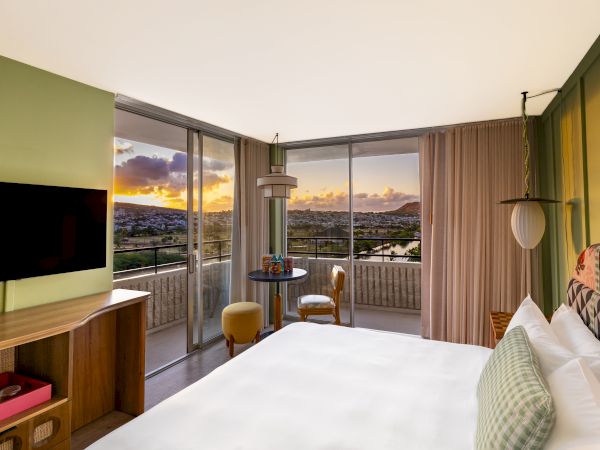 A modern hotel room with a balcony, a king bed, wall-mounted TV, and a view of the city at sunset.