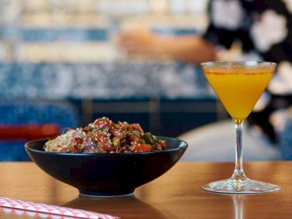 A bowl of food, possibly rice with toppings, is next to a yellow cocktail on a wooden table with a red-striped straw beside it.