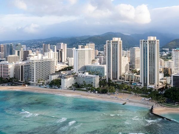 The image shows a coastal cityscape with tall modern buildings, a sandy beach, and turquoise ocean waters, set against a backdrop of mountains and cloudy skies.
