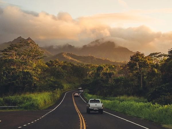 A scenic road with a vehicle driving through a lush, mountainous landscape surrounded by greenery and enveloped in soft, golden sunlight and clouds.