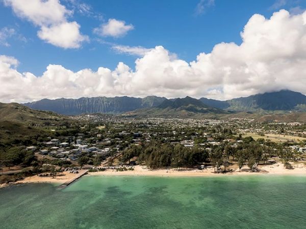 A coastal landscape with a sandy beach and turquoise waters, bordered by a residential area and mountains under a partly cloudy sky.