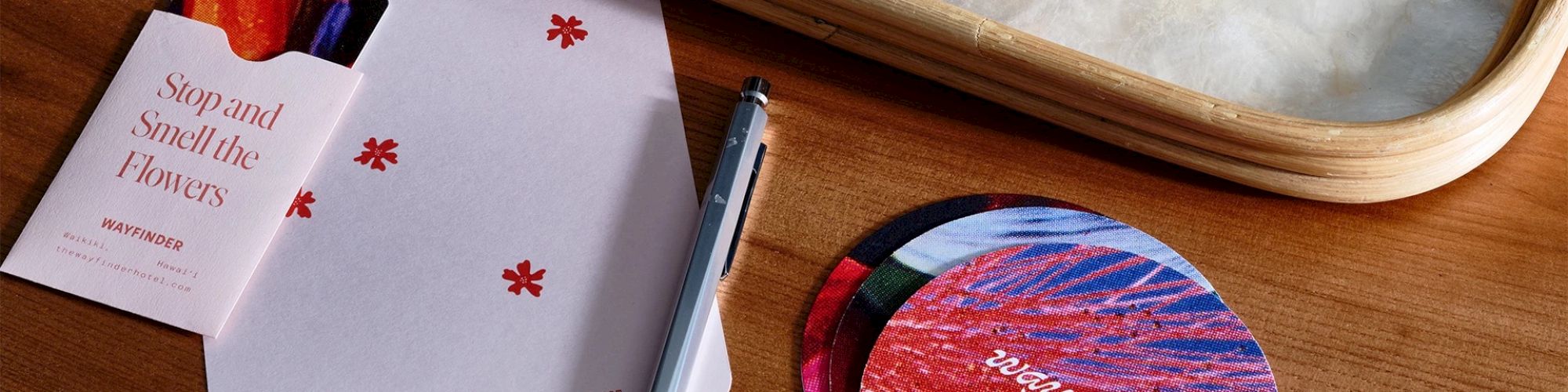 A minimalistic card reading "Stop and Smell the Flowers," a pen, circular colorful coasters, and an envelope on a wooden table.