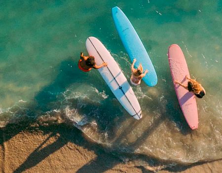 Three surfers holding surfboards&mdash;pink, blue, and white&mdash;stand at the shoreline, with clear turquoise waves gently washing over their feet.