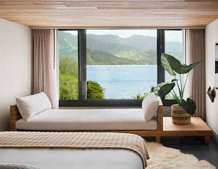 A cozy bedroom with a large window offering a scenic mountain and water view, a bench with cushions, potted plant, wall art, and wooden ceiling.