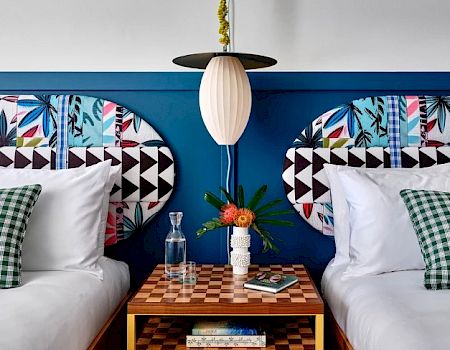 A stylish bedroom features two beds with vibrant headboards, a central hanging lamp, a checkered bedside table with books, flowers, and a water carafe.