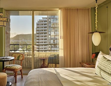 A cozy bedroom with a large bed, patterned headboard, round table, and chairs. Balcony outside with a scenic view of mountains and buildings.