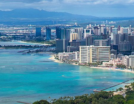 A coastal cityscape with numerous high-rise buildings, boats on turquoise waters, and mountains in the background under a partly cloudy sky.