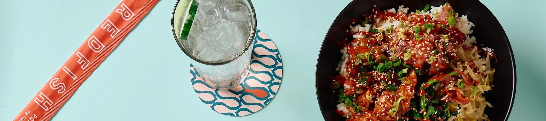 A bowl of poke with rice, vegetables, and sesame seeds, accompanied by a glass of iced drink with a lime slice and a packet labeled "REDFISH."
