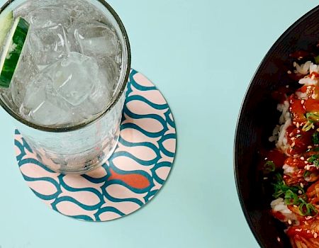 A bowl of poke with rice, vegetables, and sesame seeds, accompanied by a glass of iced drink with a lime slice and a packet labeled "REDFISH."