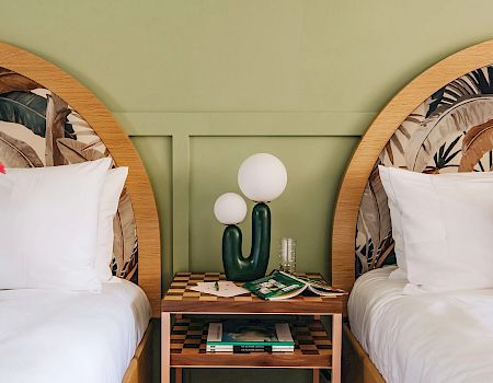 This image shows two beds with patterned headboards, green and white checkered pillows, and a nightstand with books, glasses, and a unique lamp.