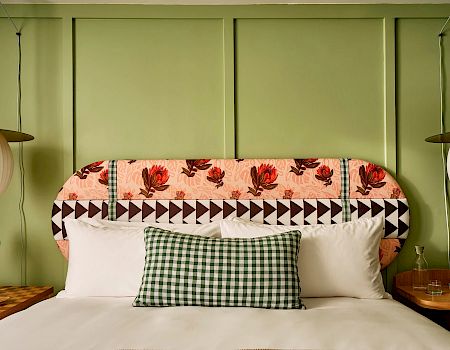 A neatly made bed features a floral-patterned headboard and gingham pillow, flanked by hanging pendant lights and nightstands with small decor items.