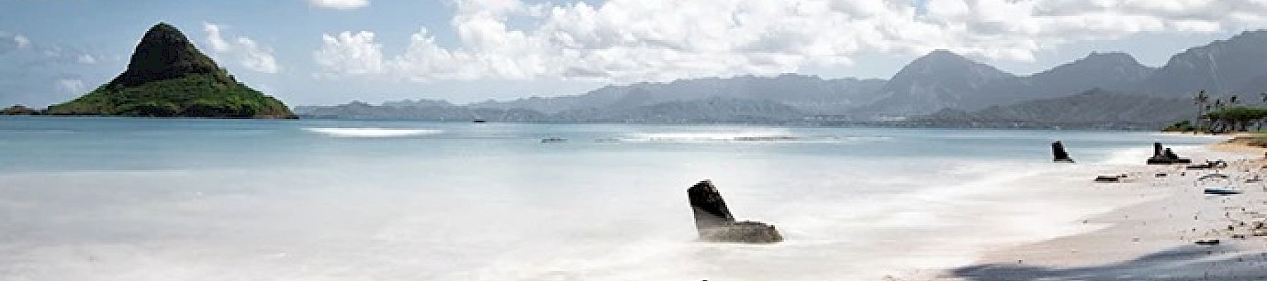 A serene beach scene with calm waters, distant mountains, a small island, and driftwood scattered along the shoreline under a partly cloudy sky.