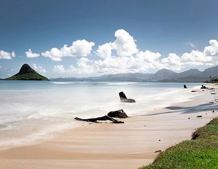 A serene beach scene with calm waters, distant mountains, a small island, and driftwood scattered along the shoreline under a partly cloudy sky.