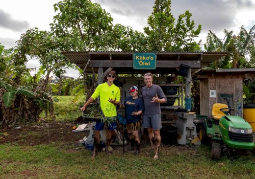 People pose at a lush roadside coffee stand under big trees with a green sign reading &ldquo;Kāko&rsquo;o ʻOiwi.&rdquo;