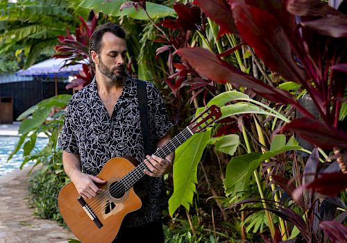 A man in a patterned shirt plays an acoustic guitar among tropical plants by a pool, creating a relaxed, tropical vibe.