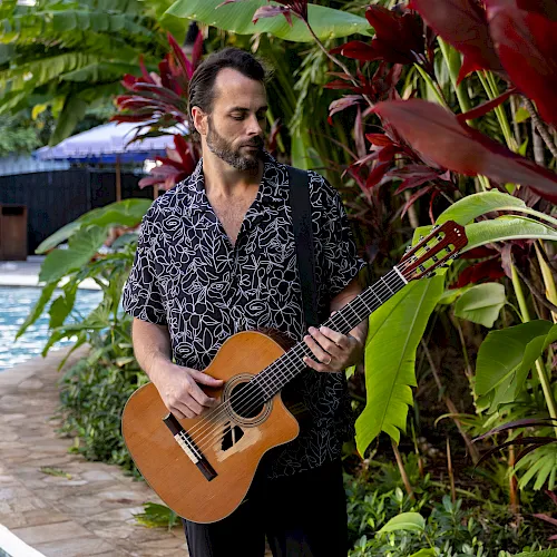 Un hombre con una camisa estampada toca la guitarra ac&uacute;stica entre plantas tropicales junto a una piscina, creando un ambiente relajado y tropical.