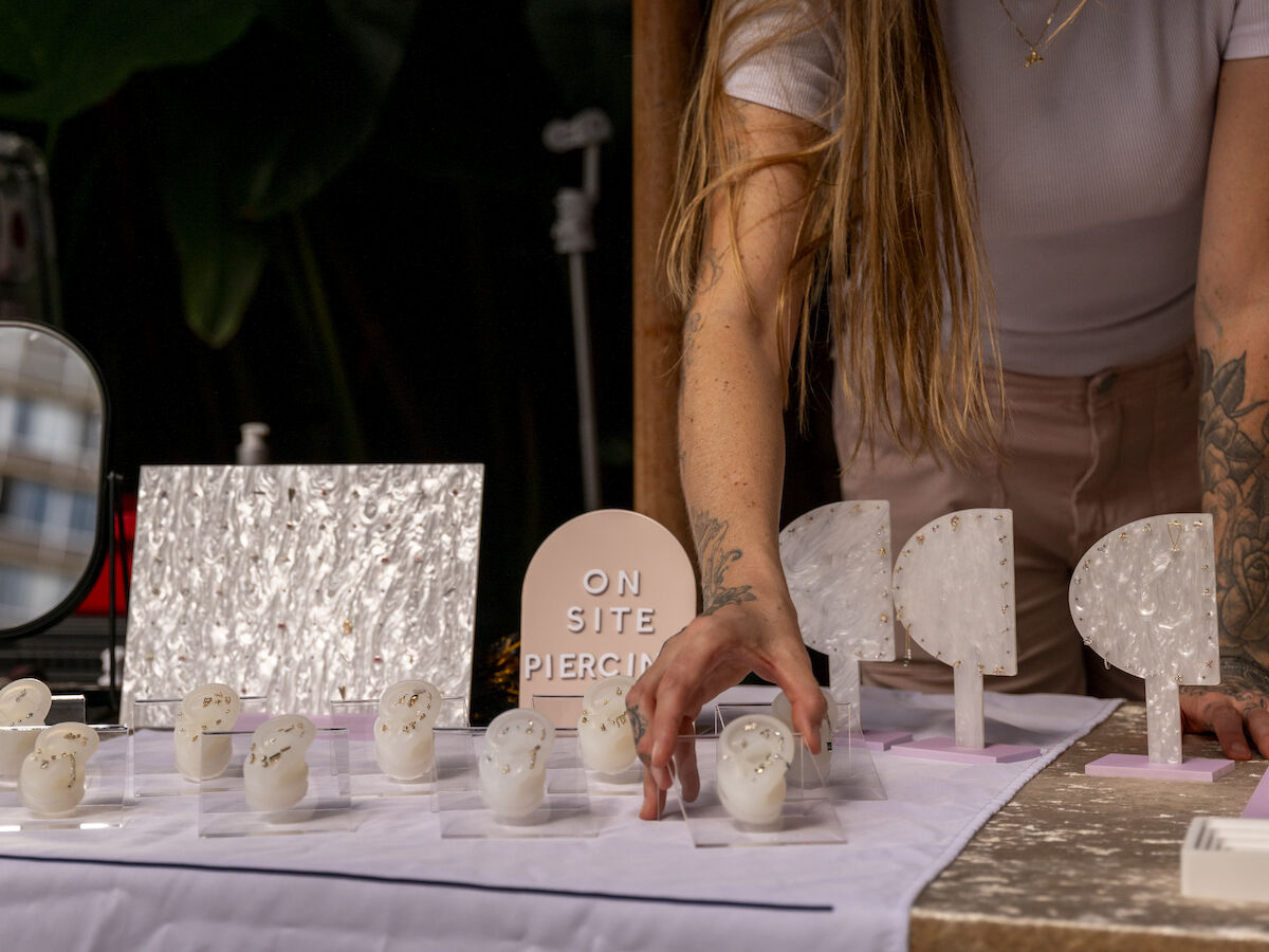 A person with tattoos sets up a display of earring stands, jewelry, and a sign that says "ON SITE PIERCING" on a table covered with a white cloth.