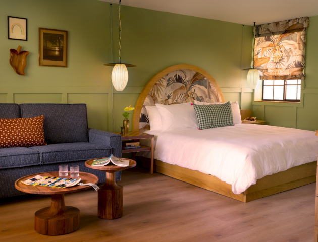 Una acogedora habitaci&oacute;n de hotel con cama king size, paredes verdes, un sof&aacute; azul, mesas r&uacute;sticas de madera, plantas y l&aacute;mparas colgantes c&aacute;lidas, que crean un refugio acogedor.