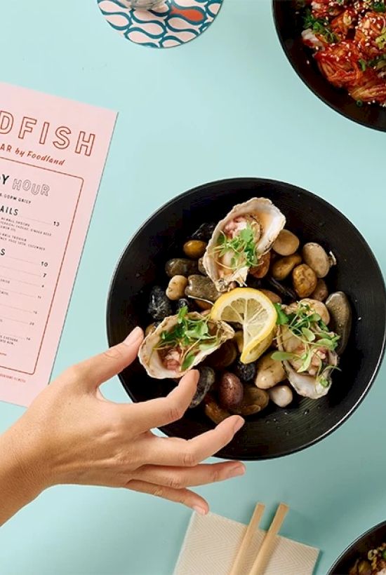 A hand reaches for a black bowl with oysters and lemon on rocks; other dishes and a "Redfish" menu also visible on a light blue table.
