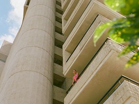 A tall building with balconies and a cylindrical structure, with a person in pink standing on one of the balconies, and some green leaves in the foreground.