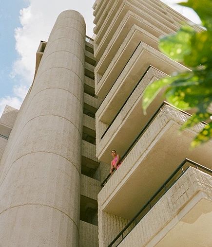 A tall building with balconies and a cylindrical structure, with a person in pink standing on one of the balconies, and some green leaves in the foreground.