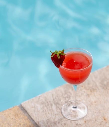 A glass of red drink garnished with a strawberry on the rim, placed on the edge of a pool.