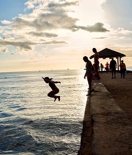 People are gathered on a pier as one person jumps into the water, with the sun setting in the background and a few clouds in the sky.