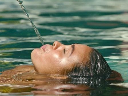 A person is floating in a pool with their head tilted back, spouting a stream of water from their mouth.