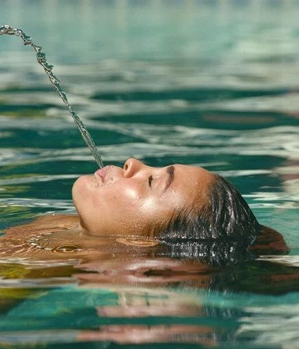 A person is floating in a pool with their head tilted back, spouting a stream of water from their mouth.
