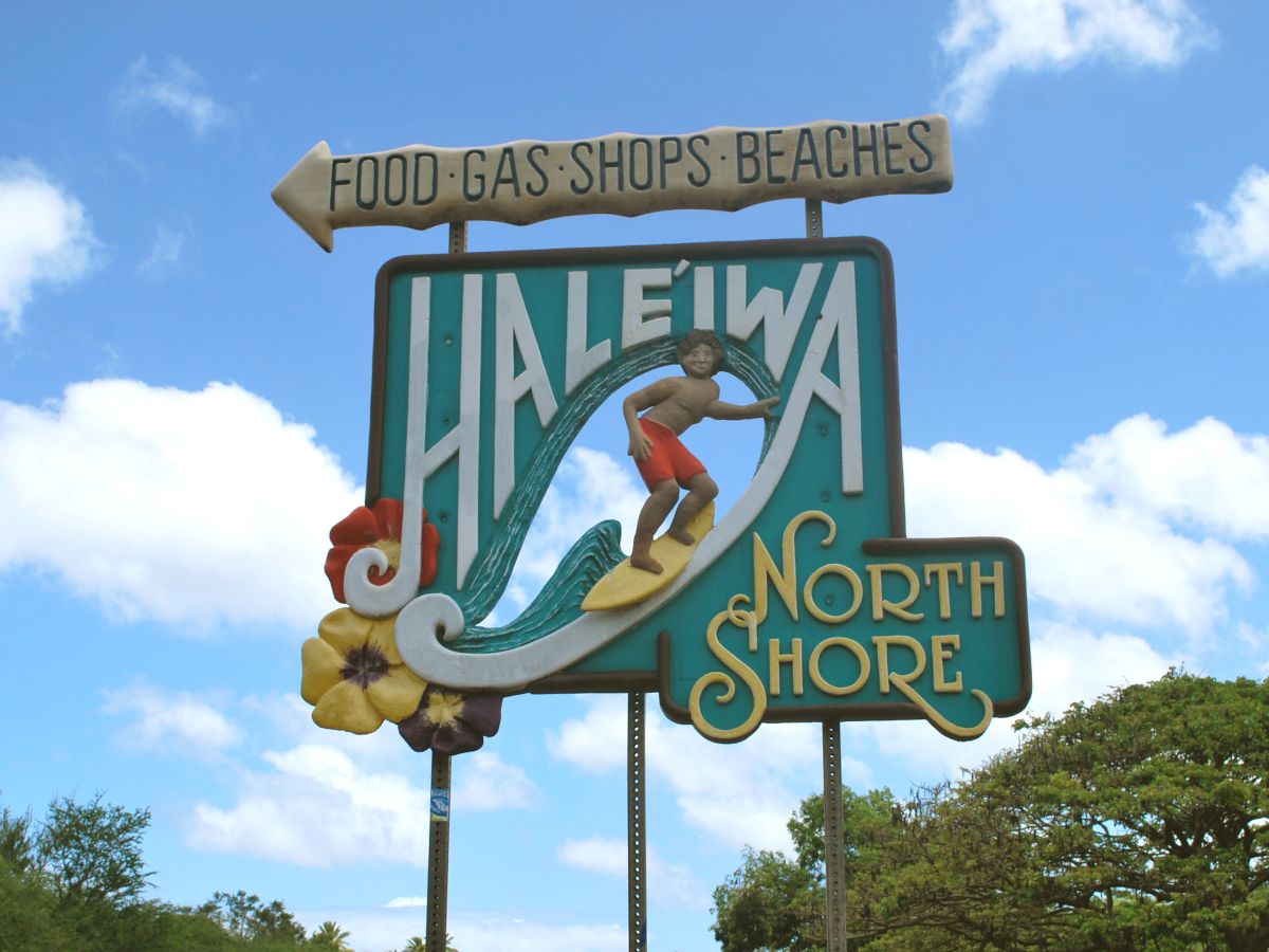 A vintage Halekala/Haleiwa sign on the North Shore, Hawaii, featuring a surfer on a wave with &ldquo;FOOD GAS SHOPS BEACHES&rdquo; top banner.