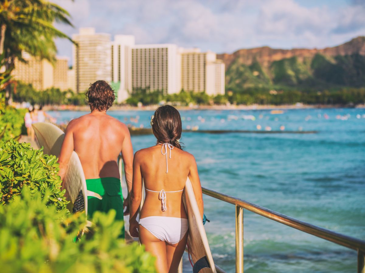 Two people in swimsuits walk along a seaside path toward the water, with buildings and mountains in the background. End with a period.