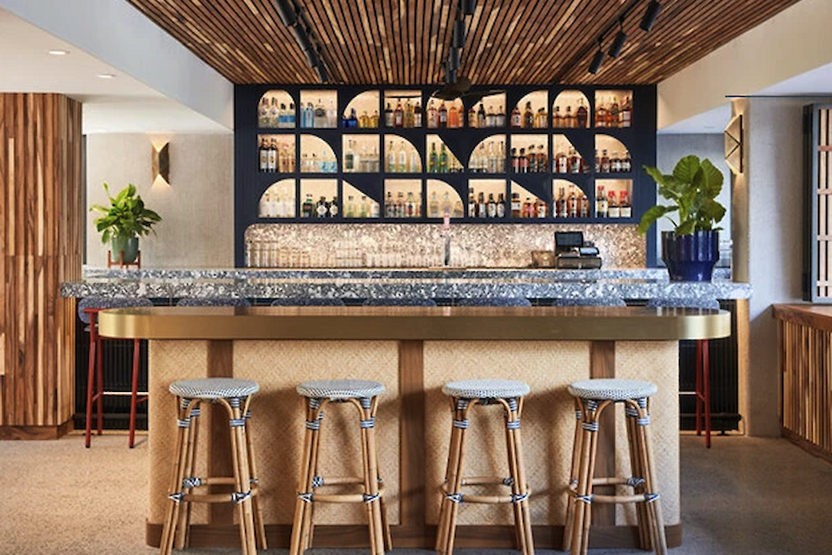 The image shows a modern bar area with a marble countertop, four bar stools, plants, and a stylish back wall with shelves displaying bottles.