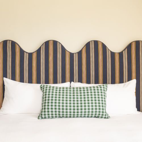 A bed with white pillows, a green checkered cushion, striped headboard, and blue hanging lamps. There&rsquo;s a small table with plants and decor.