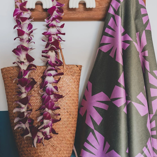 A woven basket with a purple lei, a wooden wall hook, and a gray towel with pink patterns hang on a blue and white wall.