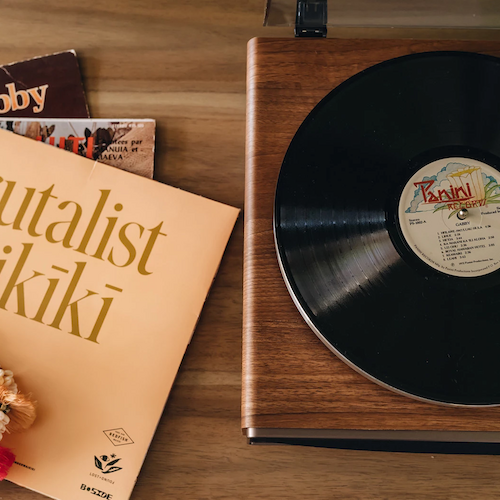 A vinyl record is spinning on a wooden turntable. To the side are various record covers, including "Brutalist Waikiki," alongside seashell necklaces.
