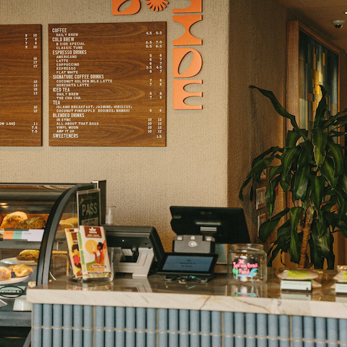 This image shows a caf&eacute; counter with a cash register, a plant, a menu on the wall, and a glass display case with food items.