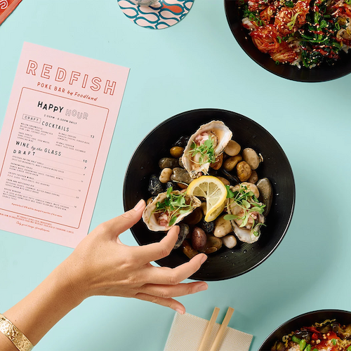 A person reaches for a bowl of oysters with lemon slices and garnishes; nearby are sushi bowls, a menu with "REDFISH," a patterned bowl, and chopsticks.