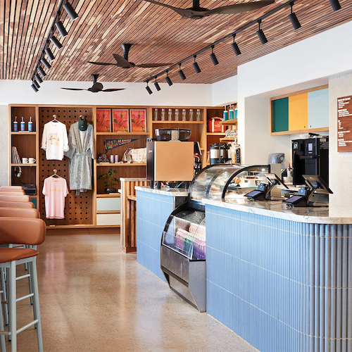 A modern caf&eacute; with a counter display, bar stools, ceiling fans, and a retail area showcasing clothing and other items on shelves.