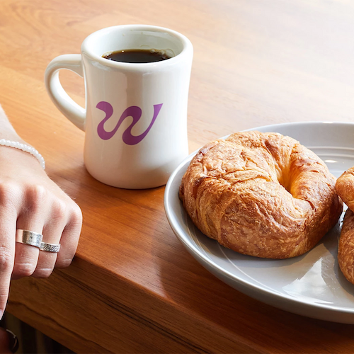 The image shows a hand wearing rings and a bracelet, a white coffee mug with a purple "W" logo, and a plate with two croissants on a wooden table.